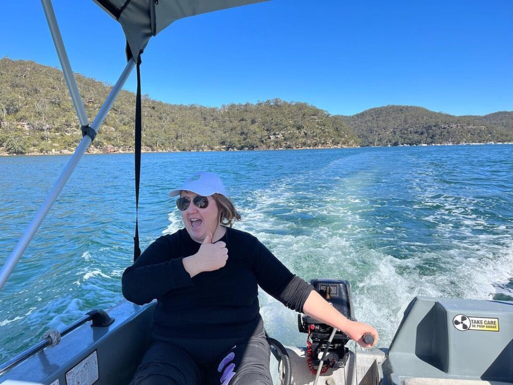 Rahni Allan in a white hat and sunglasses steering a boat, laughing joyously, with a forested coastline in the background.