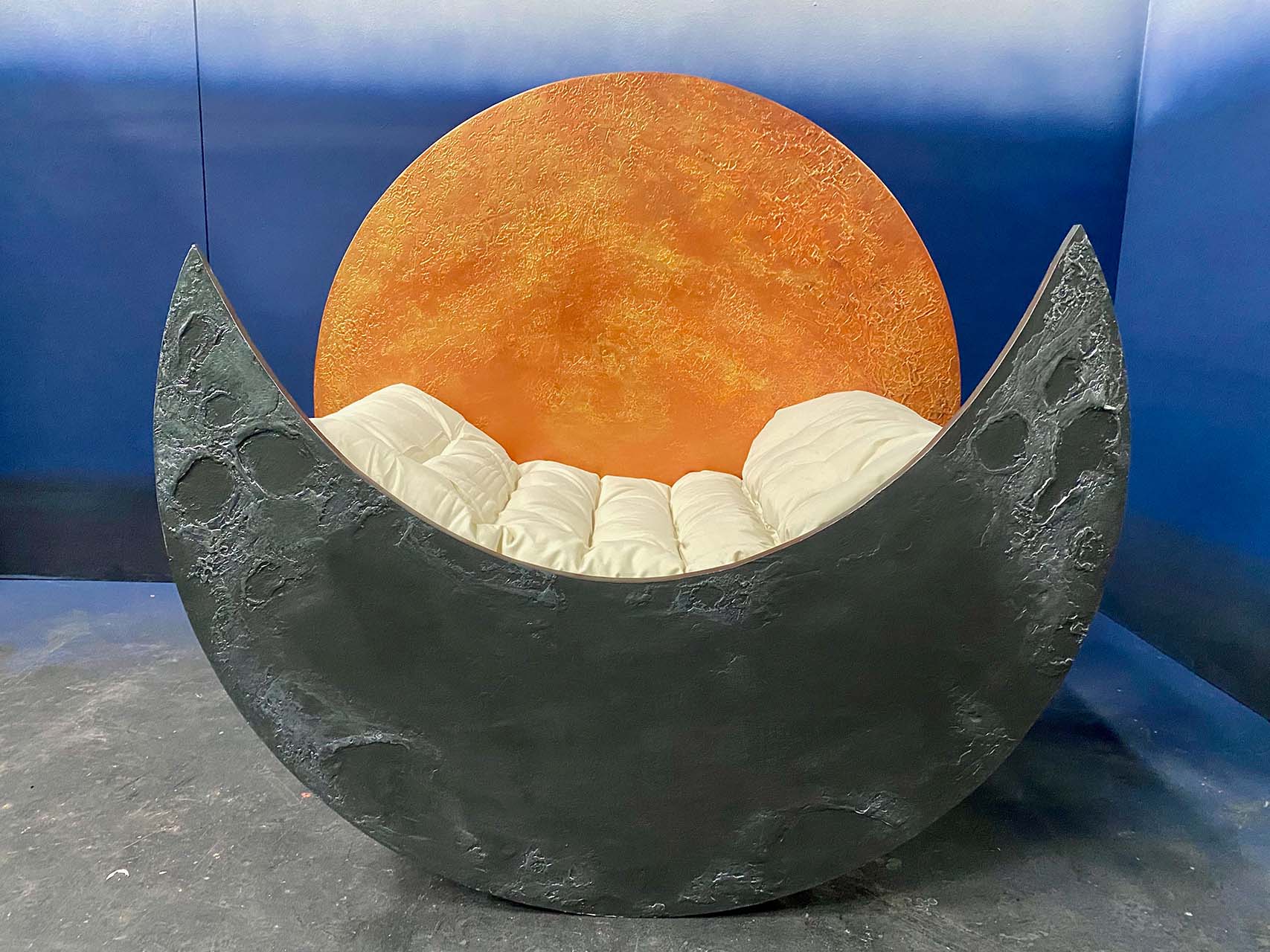 A creative chair designed to resemble a crescent moon, with a large orange spherical backdrop and white cushions placed inside the chair.