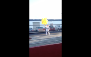 A person in white clothing holds a bunch of yellow balloons while standing in a parking lot with several cars and a white building in the background.