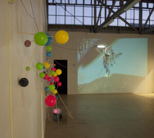Colorful balloons and sticks form a wall-mounted sculpture in an art gallery; a projection of a landscape with an abstract structure is displayed on the adjacent wall.