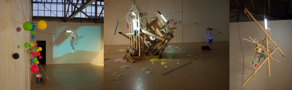 Three-panel view of an art installation with colorful balloons, wooden structures, lights, projected image, and scattered materials on a gallery floor.