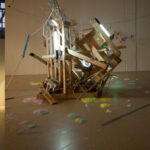 Three-panel view of an art installation with colorful balloons, wooden structures, lights, projected image, and scattered materials on a gallery floor.