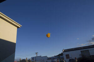 A cluster of yellow balloons floats in a blue sky above industrial buildings, with power lines and rooftops visible below.