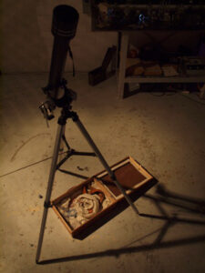 A telescope on a tripod stands on a concrete floor, with an open wooden case containing various items placed beneath it.