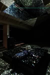 A person lies on a dark platform in a dim room, illuminated by scattered star-like light projections from above and around.