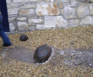 A large dark rock is partially embedded in gravel near a stone wall with a vent. Another smaller rock and a person’s leg are also visible.