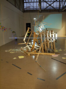 Wooden planks, fluorescent lights, and ladders are arranged in a sculptural installation in a large, industrial gallery space with scattered debris on the floor.