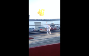 A person in white clothes stands in a parking lot, reaching up toward a cluster of yellow balloons floating away above parked cars.