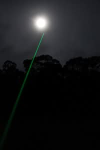 A green laser beam points upward toward a bright full moon in a dark night sky, with tree silhouettes visible below.