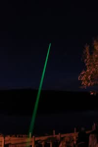 A green laser beam points upward into the night sky from the ground near a body of water, with trees and a fence visible in the foreground.