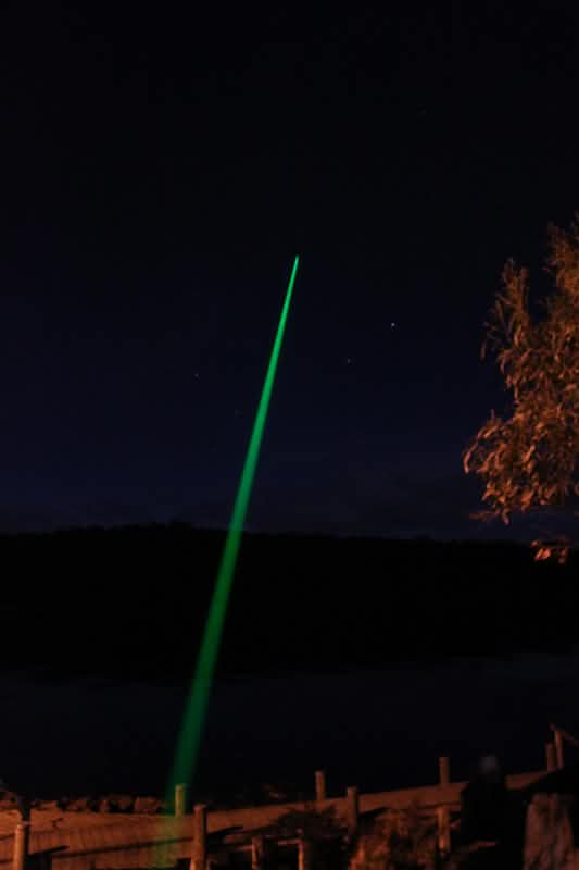 A green laser beam points upward into the night sky from the ground near a body of water, with trees and a fence visible in the foreground.