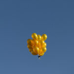 A cluster of yellow balloons floats high in a clear blue sky.