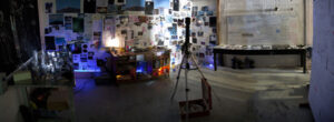 A dimly lit room with photo-covered walls, a workbench with tools, a tripod, and a table with papers, resembling a creative workspace or investigation setup.