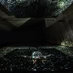 A person lies on the floor in a dark room, surrounded by scattered light projections resembling stars or galaxies on the walls, ceiling, and floor.