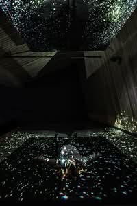 A person lies on the floor in a dark room, surrounded by scattered light projections resembling stars or galaxies on the walls, ceiling, and floor.