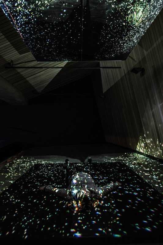 A person lies on the floor in a dark room, surrounded by scattered light projections resembling stars or galaxies on the walls, ceiling, and floor.