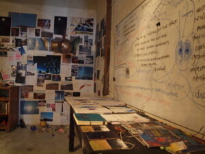 A cluttered room with papers, photos, and notes covering the walls, a whiteboard filled with diagrams, and a desk scattered with documents and books.