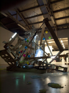 A wooden structure with colorful balloons, lights, and various objects is displayed in a gallery with a high ceiling and exposed rafters.