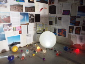 A wall covered with papers and photos, with a variety of balls and a large glowing sphere arranged on the floor in front.