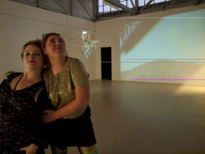 Two women stand together in an art gallery with a projection of a mountain landscape displayed on the wall behind them.