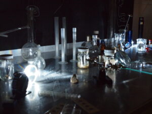 A dimly lit laboratory workspace with glassware, jars, graduated cylinders, and small scientific instruments on a metal surface.