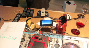 A cluttered table with audio equipment, remote controls, a smartphone, a purse, papers, and two glasses of red wine.