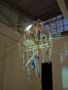 A suspended wooden sculpture made of chairs and sticks is hanging from the ceiling in a dimly lit industrial space.